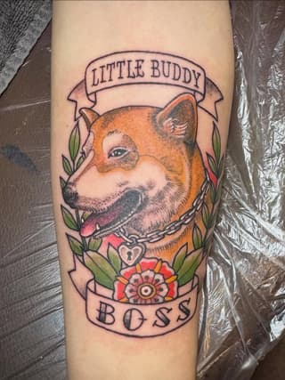 Colorful dog portrait tattoo with "Little Buddy" banner above and "Boss" banner below, surrounded by green leaves and red flowers on the forearm.