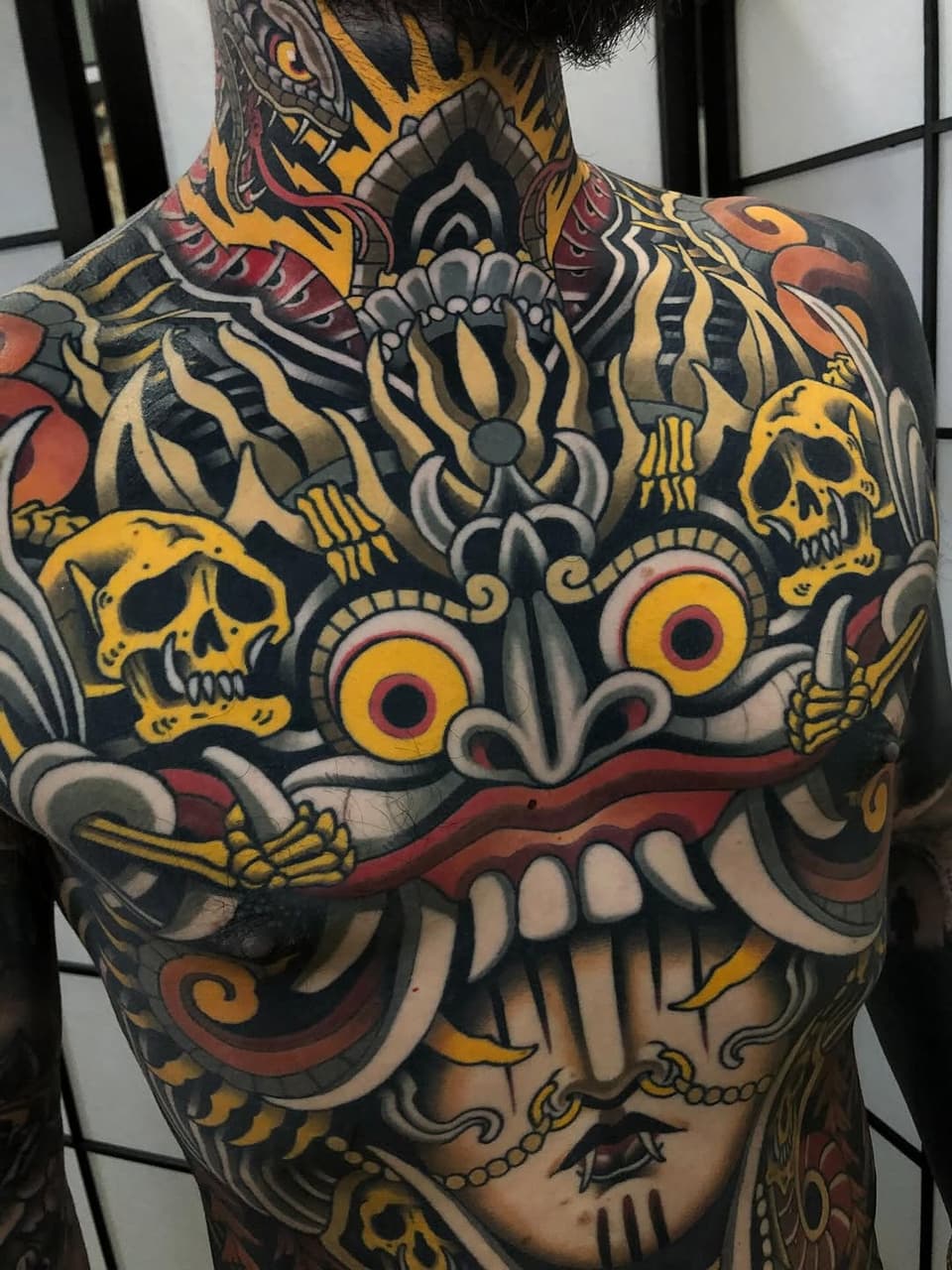 Large colorful back tattoo featuring skulls, ornamental flowers, and decorative elements in yellow, red, black, and gold tones.