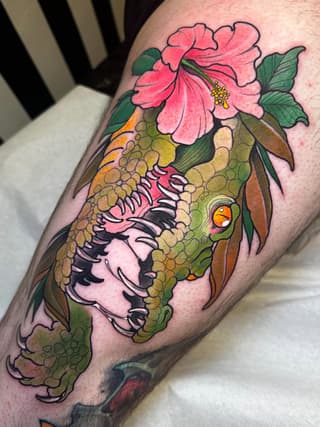 A colorful tattoo on the upper arm featuring a reptilian head with scaled texture, pink flower, and green leaves in shades of green, pink, orange, and yellow.