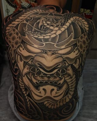 Large back tattoo featuring a multi-faced demon mask design with intricate scale patterns, shading, and ornamental details in black and tan ink.