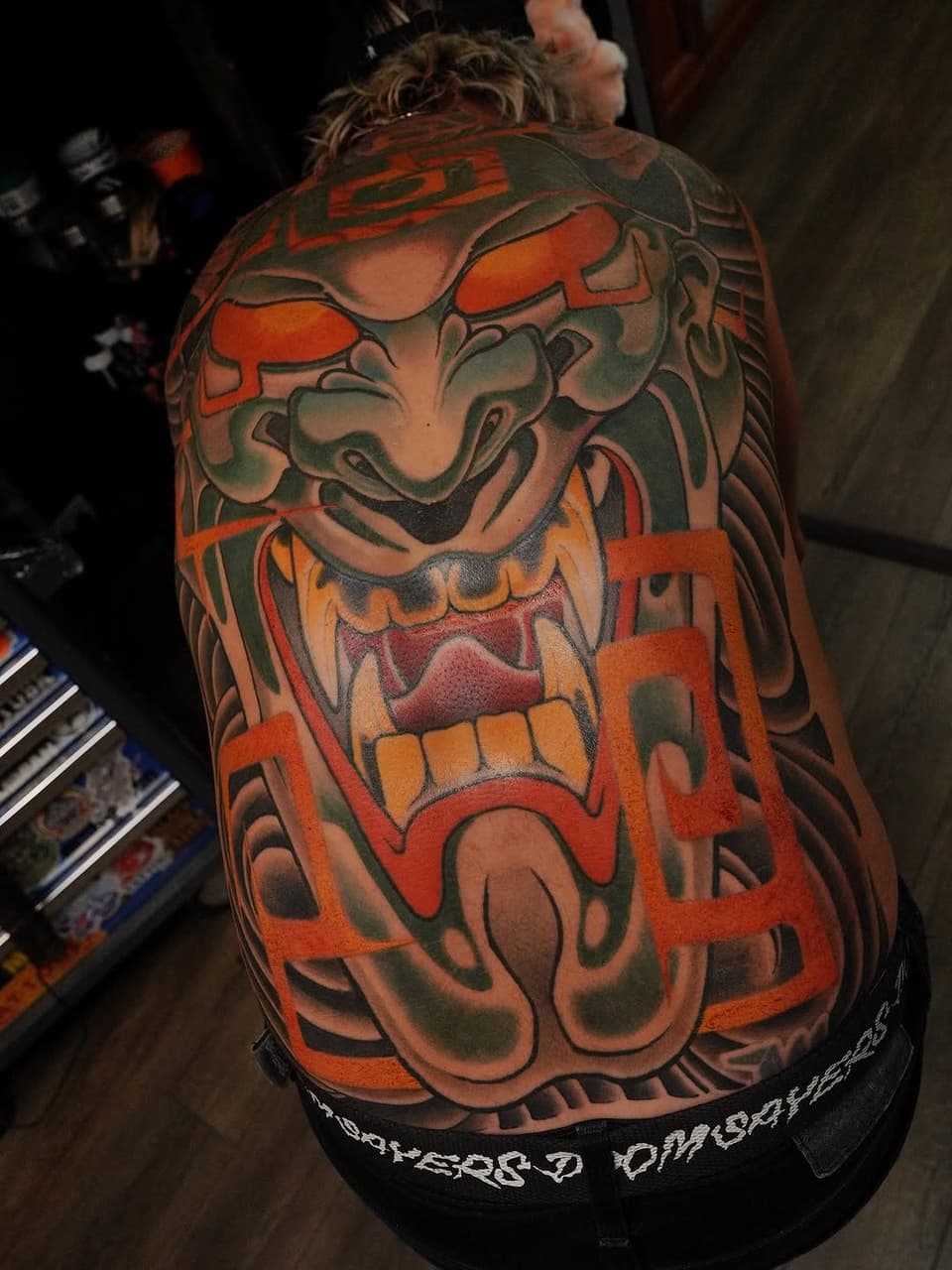 Large colorful demon or oni mask tattoo on upper back featuring orange, red, and green tones with bold outlines and shading.