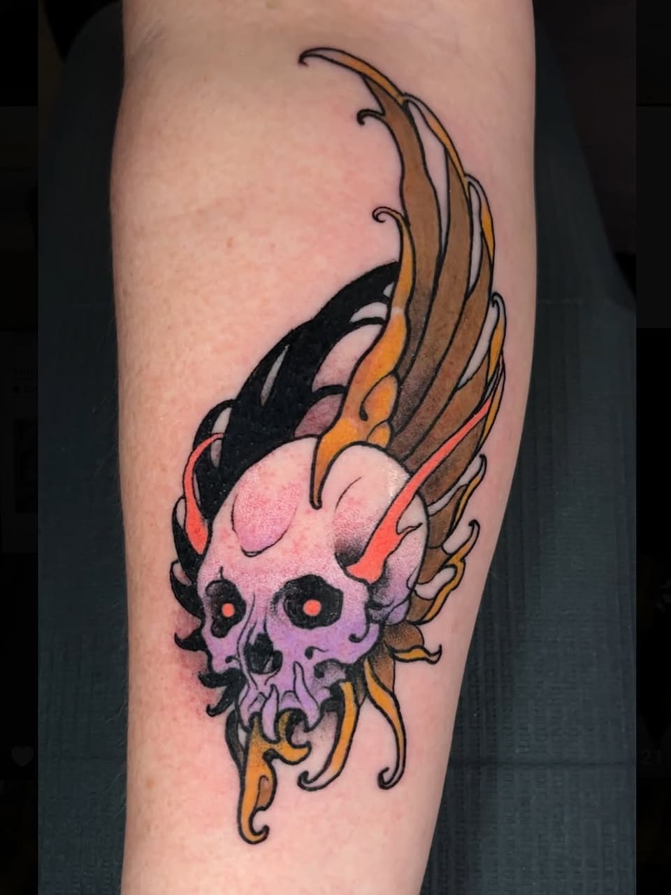 Colorful skull tattoo with flaming wings in purple, orange, and black on a forearm, featuring glowing red eyes and decorative swirls.