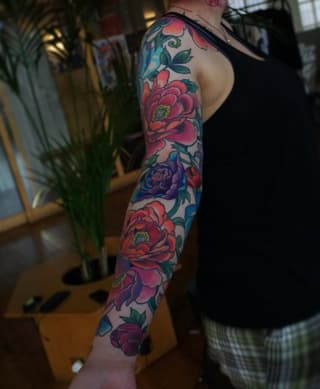 Full-length arm sleeve tattoo featuring multiple red and purple flowers with green leaves and blue accents on a person's right arm.