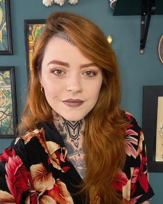 Woman with reddish-brown hair displaying a geometric chest and neck tattoo in black ink with symmetrical line patterns.