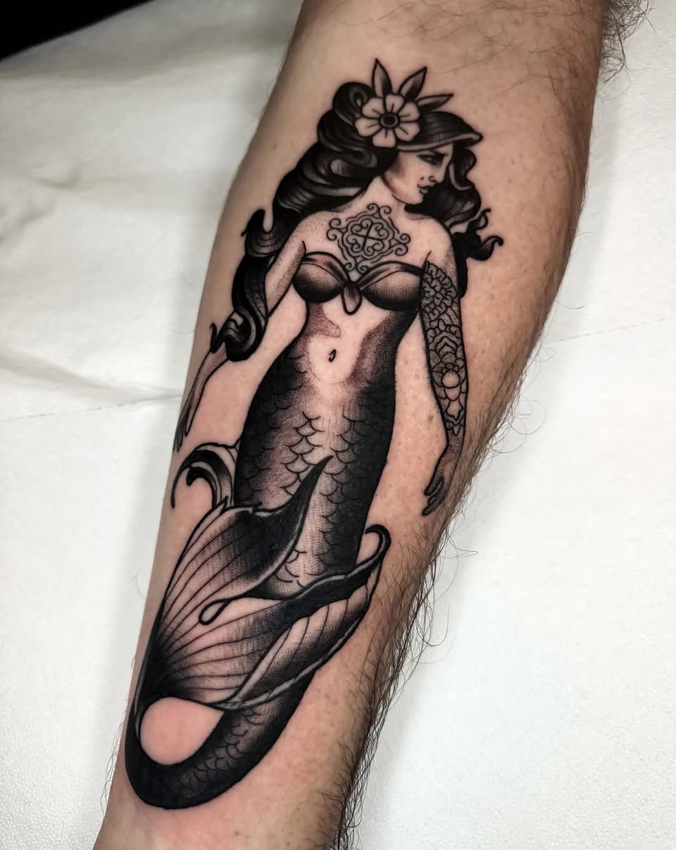 Mermaid figure with flowing hair, flower crown, and detailed tail scales tattooed on a forearm in black ink with shading.