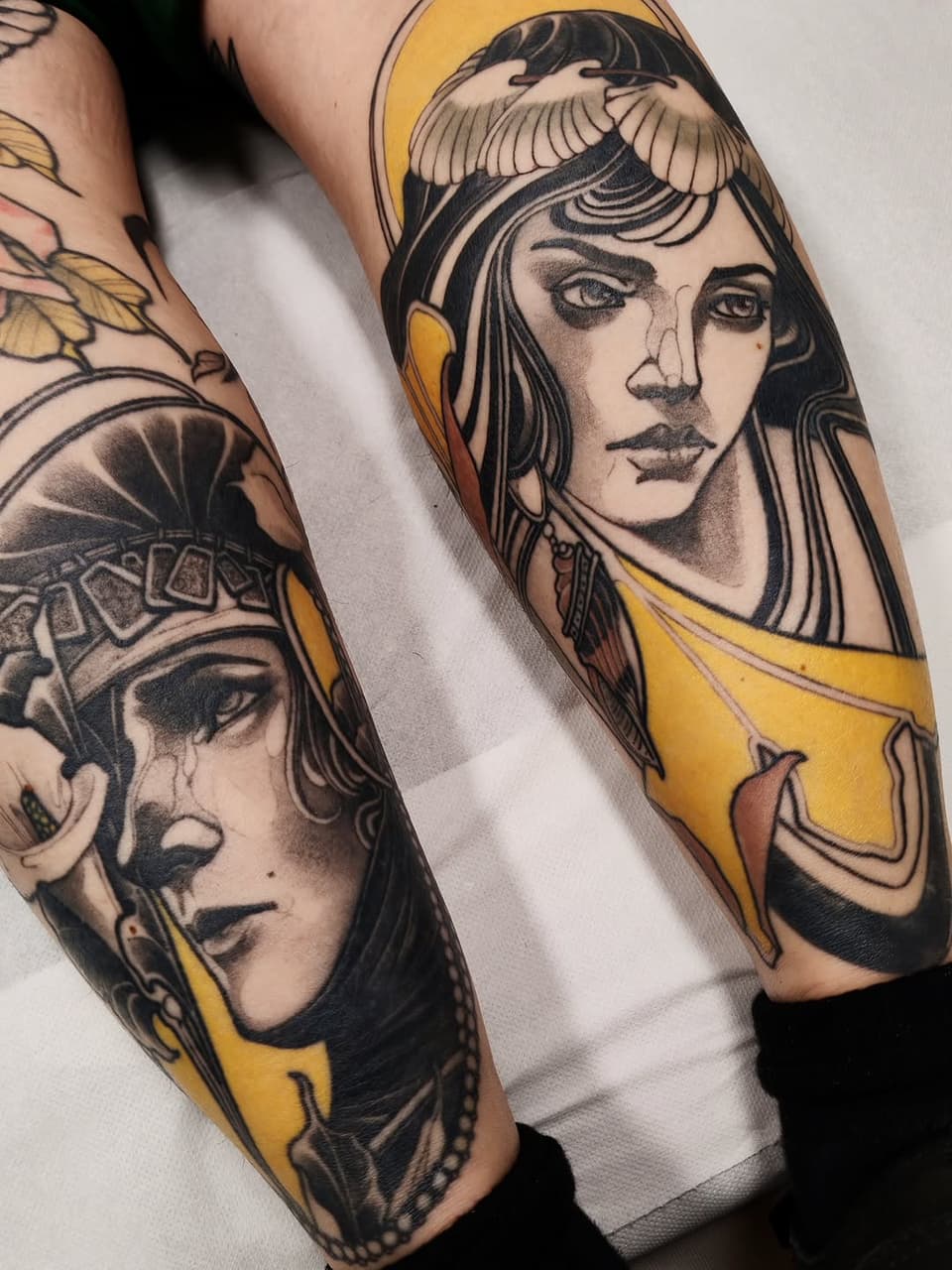 Two Egyptian-inspired female face tattoos on forearms featuring winged headdresses, gold accents, and black line work with shading details.