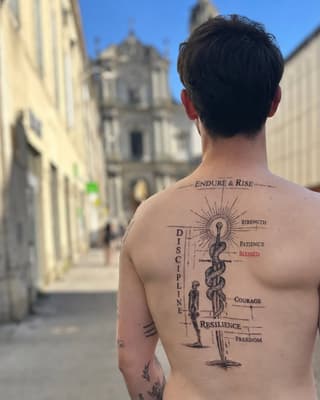 Shirtless man displaying large back tattoo featuring medical caduceus symbol, human figure, and words including "Endure & Rise," "Discipline," and virtues, set in historic stone street.