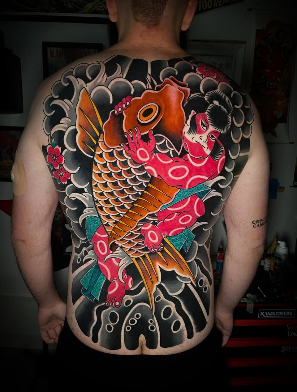 Colorful koi fish tattoo covering the entire back with orange and red scales, surrounded by black cloud motifs and small red flowers.