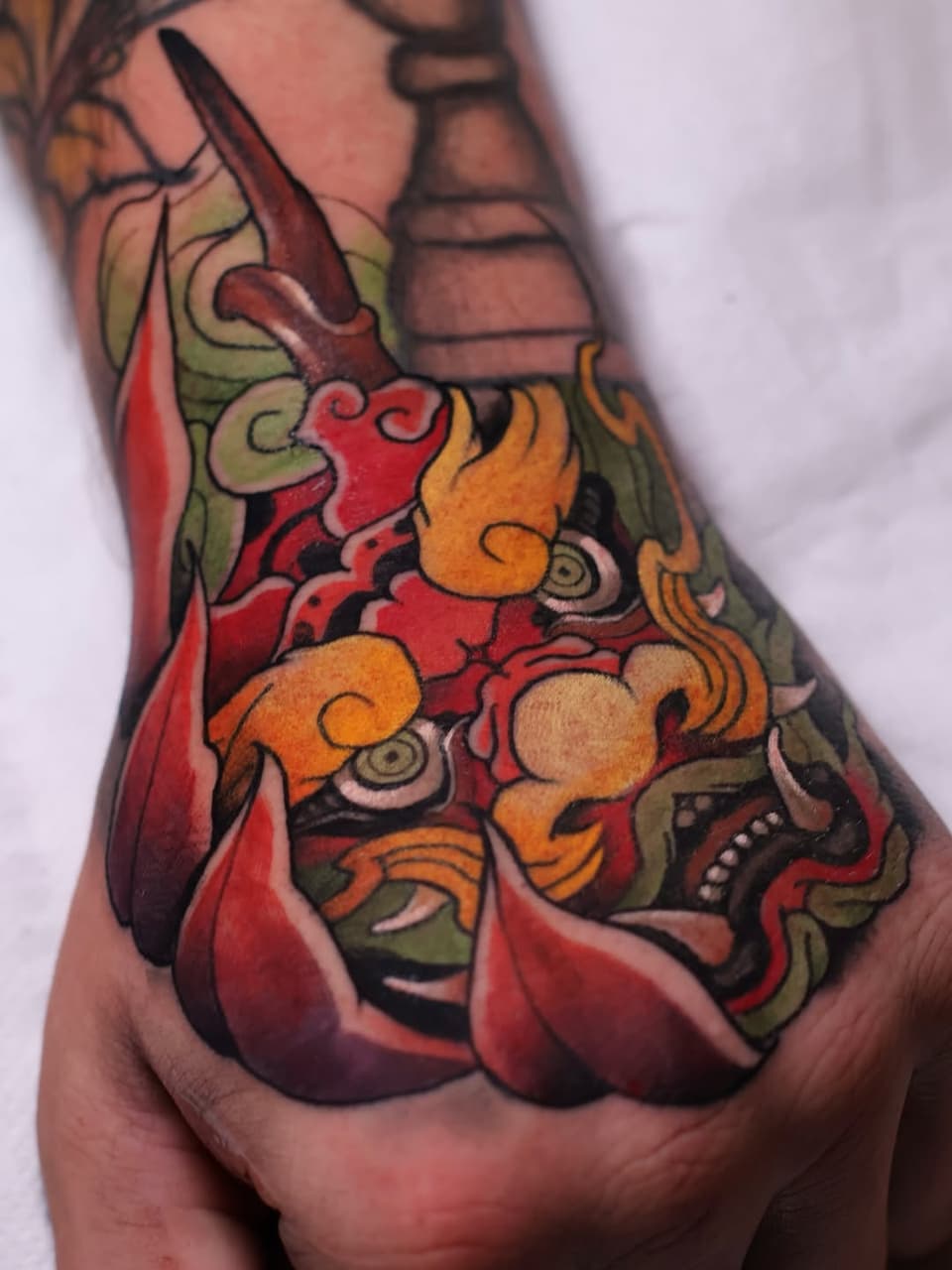 Colorful hand tattoo featuring ornamental creatures with orange, red, and green details, intricate scrollwork, and decorative elements on the knuckles and fingers.