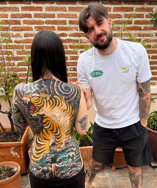Back tattoo featuring a large yellow and black striped tiger with grey wings and red accents, paired with teal and grey waves below.