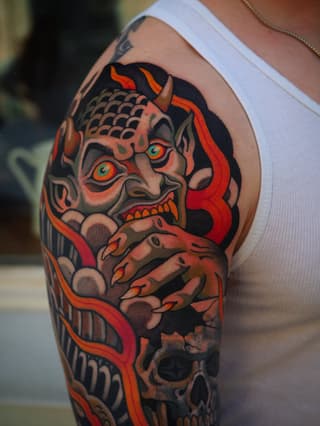 Colorful demon or oni mask tattoo on upper arm featuring red eyes, orange and black details, with a skull visible below, displayed on pale skin.
