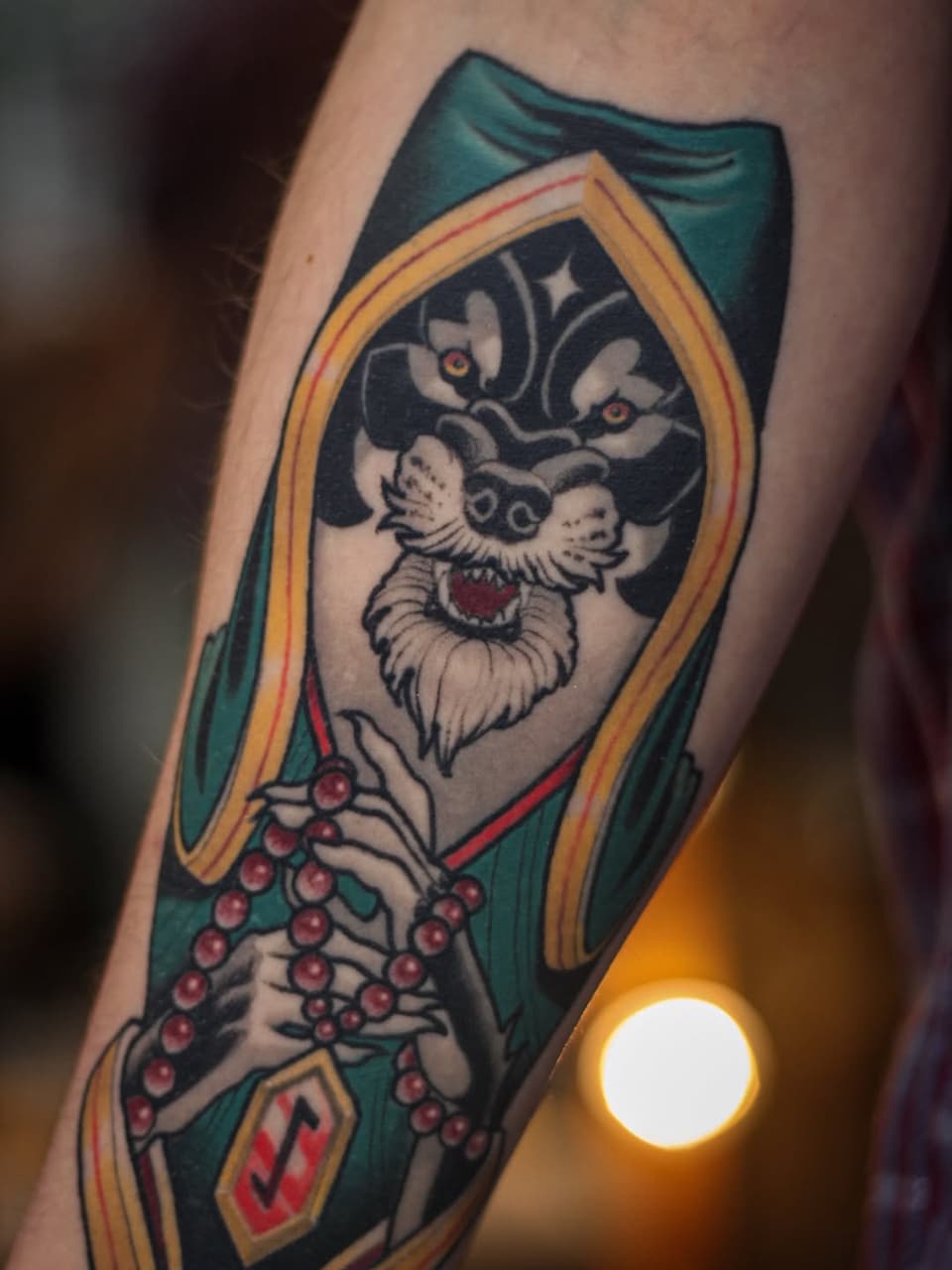 Coffin-shaped tattoo featuring a skeletal face with red eyes, wearing a green cloak and holding red beads, framed in gold and yellow borders.