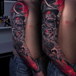 Full-sleeve tattoo featuring a demon mask with red accents, ornamental scrollwork, and decorative elements in shades of gray, red, and orange on both arms.