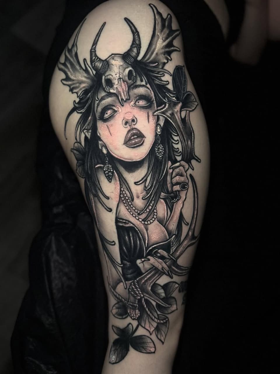 Tattoo of a demon-horned woman with flowing hair, pearl necklace, and floral elements on the upper arm in black and grey ink with skin tone highlights.