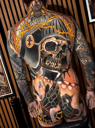 Full-back tattoo featuring a ornate skull with crown and crossbones, rendered in orange, black, and grey on human skin.