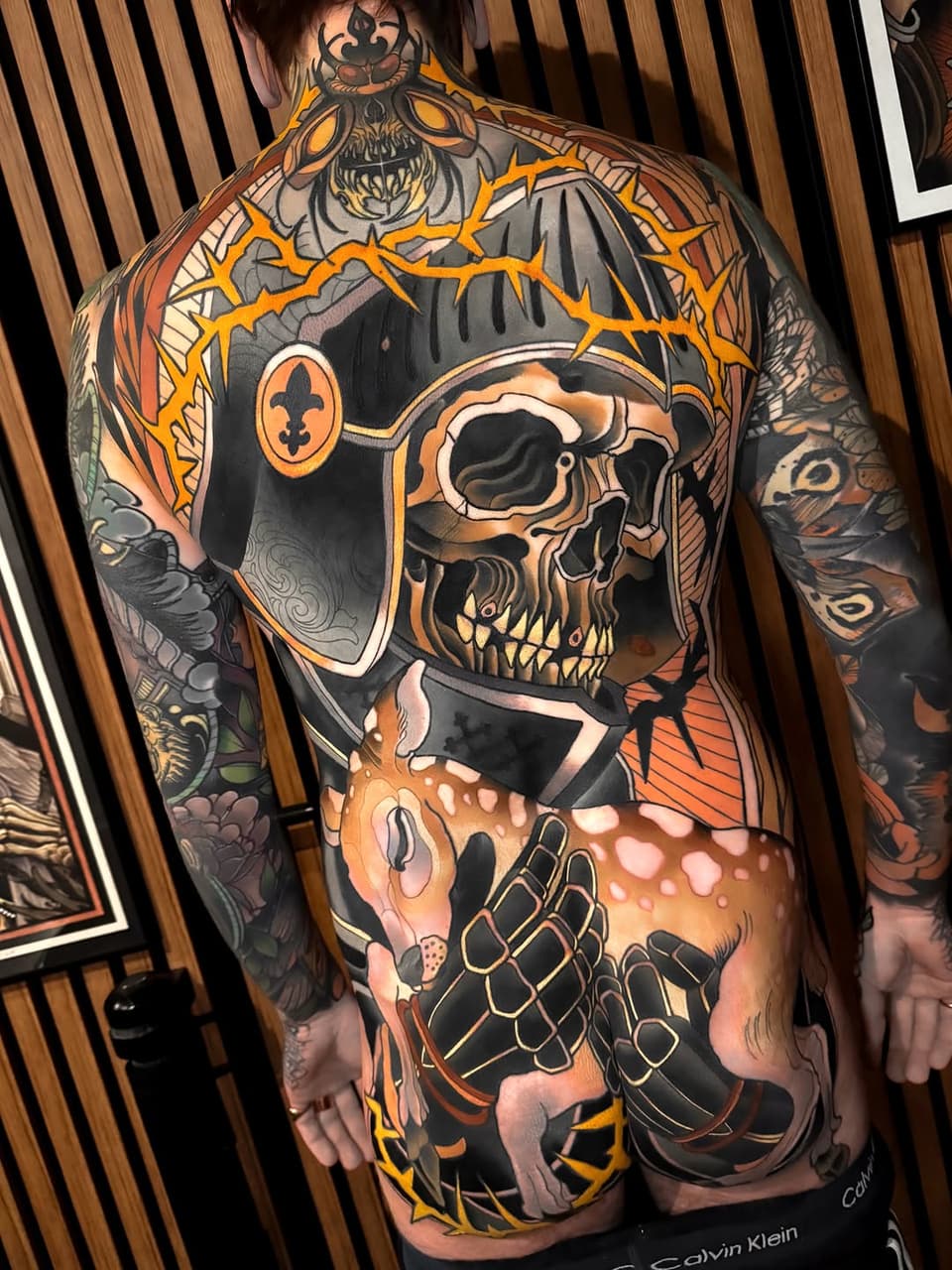 Full-back tattoo featuring a ornate skull with crown and crossbones, rendered in orange, black, and grey on human skin.