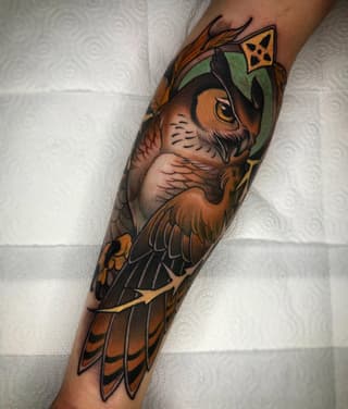 Detailed owl tattoo on a forearm featuring brown and orange plumage, green headpiece, yellow eyes, and geometric crown, displayed against a white textured background.