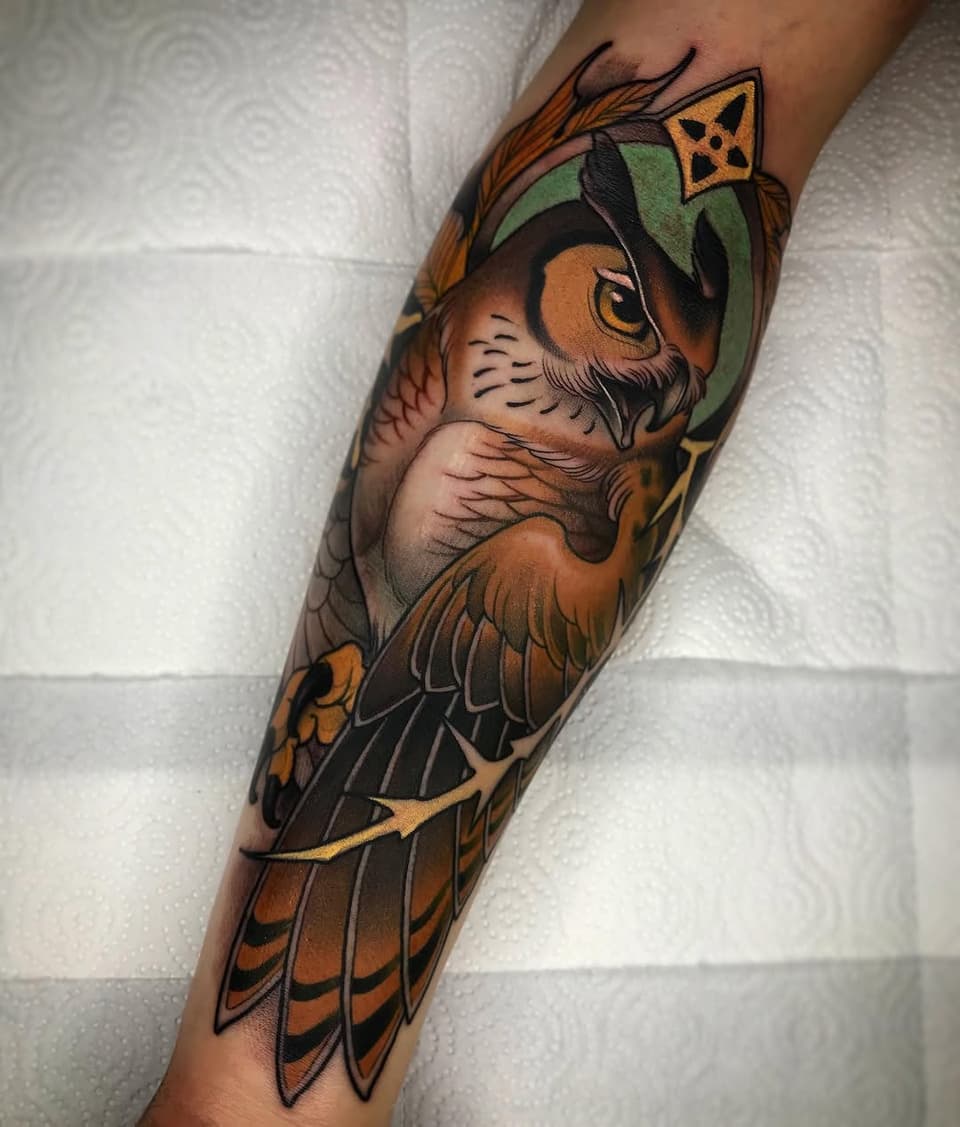 Detailed owl tattoo on a forearm featuring brown and orange plumage, green headpiece, yellow eyes, and geometric crown, displayed against a white textured background.