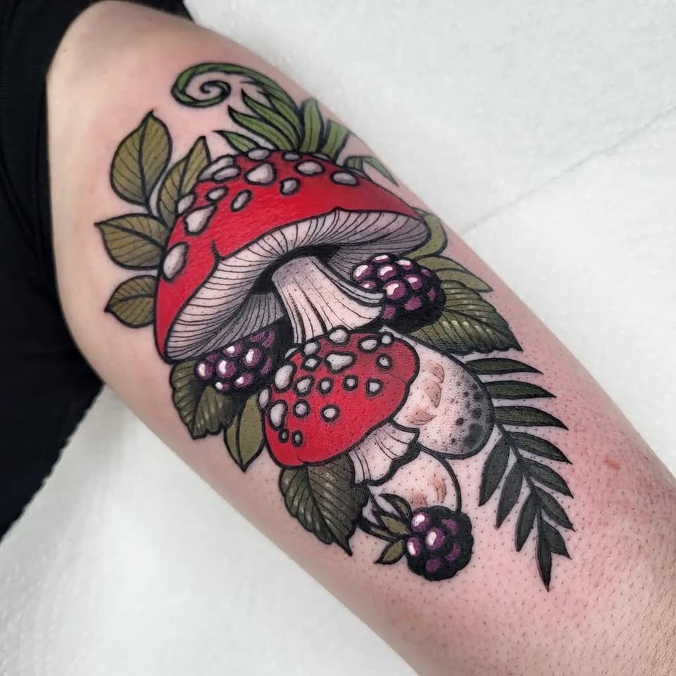 Colorful arm tattoo featuring red mushrooms with white spots, blackberries, green leaves, and a decorative swirl, arranged vertically on the upper arm.