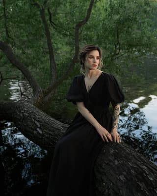 Woman wearing black dress seated on tree branch over water displays an ornate floral tattoo on her right forearm in dark ink.