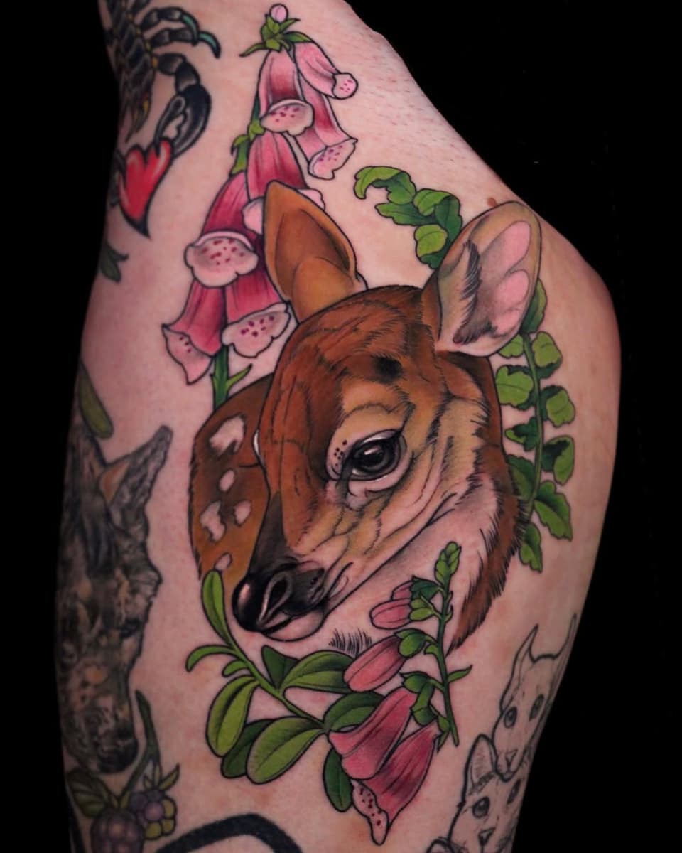 Colored tattoo on thigh of a brown deer's head surrounded by pink hanging flowers and green foliage.
