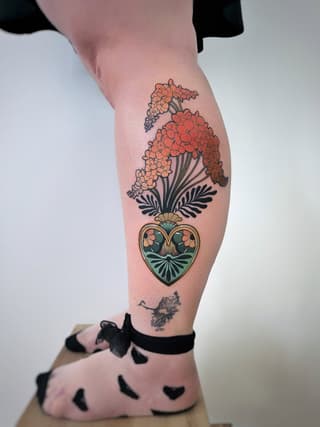 Colorful leg tattoo featuring clustered orange flowers with dark foliage sprouting from a green and gold heart-shaped vessel.