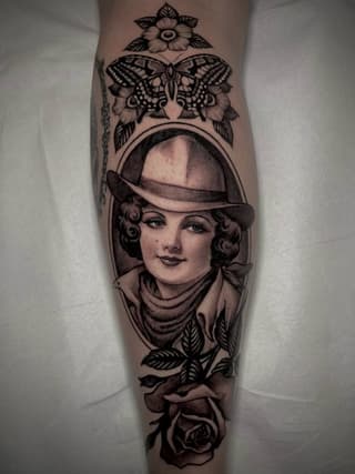 Vertical leg tattoo featuring a portrait of a woman in a fedora hat surrounded by a butterfly, flowers, and rose, rendered in black ink.