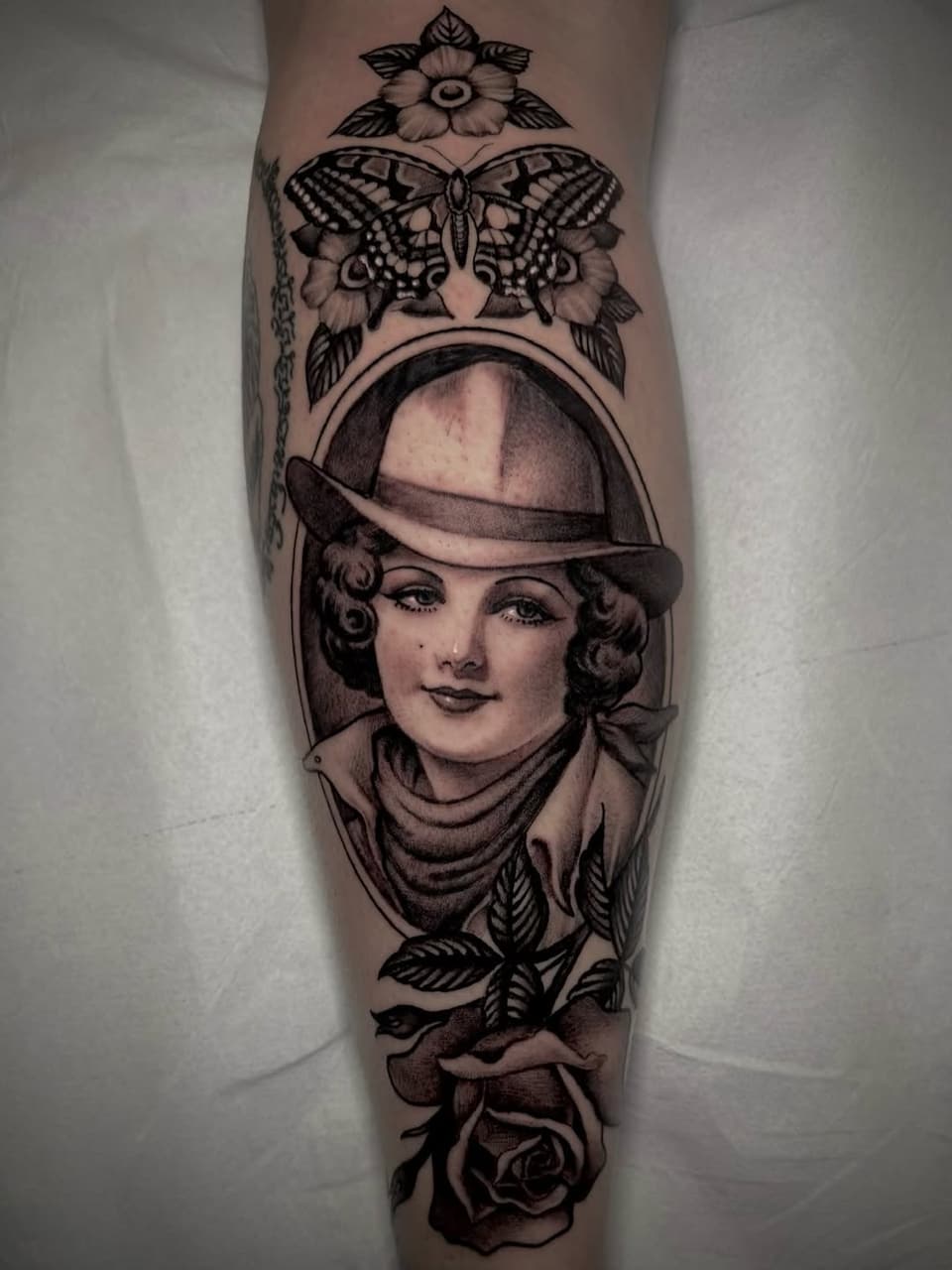 Vertical leg tattoo featuring a portrait of a woman in a fedora hat surrounded by a butterfly, flowers, and rose, rendered in black ink.