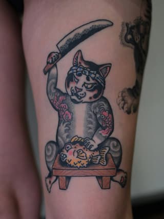Tattooed cat with decorative markings wielding a curved blade while sitting on a bench, featuring pink, orange, and black ink on thigh.