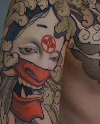 Tattoo on upper arm featuring a stylized face with golden eyes, red facial markings, ornate curled hair details in tan and black, and red mouth covering.