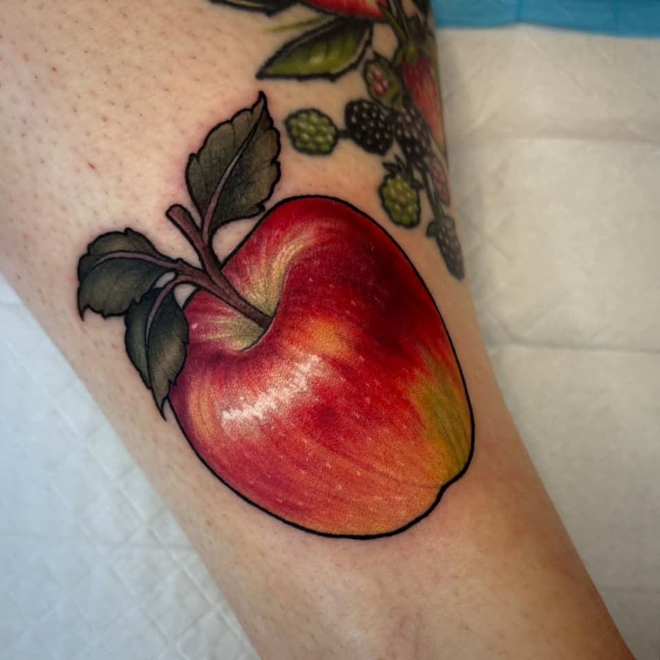 Colorful apple tattoo with green leaves on the upper arm, featuring red, orange, and yellow shading with black outlines.