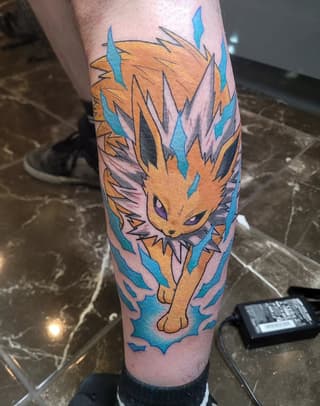 Golden cat with spiky mane and blue eyes surrounded by blue flame-like shapes, tattooed on the calf in orange, blue, pink, and black ink.