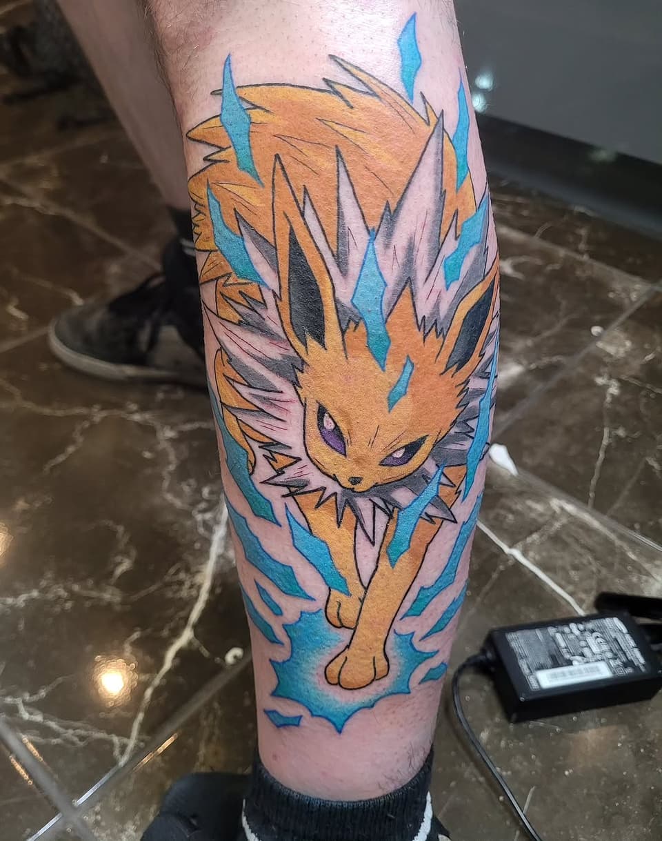 Golden cat with spiky mane and blue eyes surrounded by blue flame-like shapes, tattooed on the calf in orange, blue, pink, and black ink.
