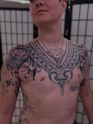 Shirtless man displaying extensive chest and shoulder tattoos featuring ornamental patterns, floral designs, and geometric motifs in black ink.
