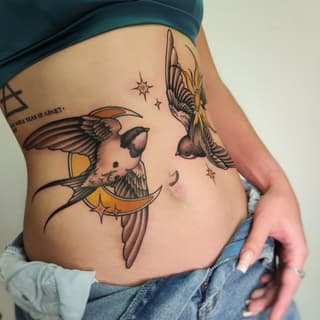Winged birds perched on crescent moons with stars and text on the upper arm and side ribs, rendered in black ink with gold and orange accents.