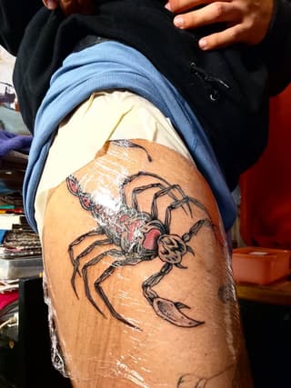 Spider and moon tattoo on upper thigh in black ink with red and skin-tone details, currently wrapped in plastic during healing.