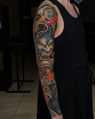 Full-sleeve arm tattoo featuring a demon mask with ornamental scrollwork, red flowers, and gold accents on a dark background.
