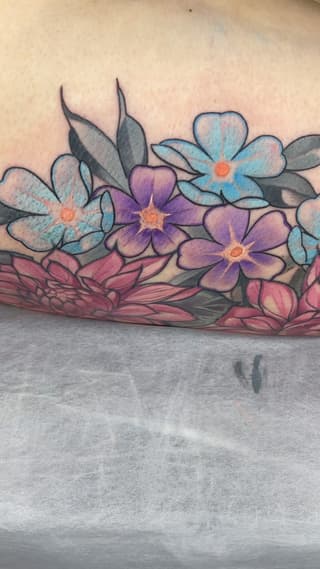 Colorful floral tattoo on forearm featuring blue, purple, and pink flowers with orange centers and green leaves arranged horizontally.