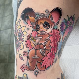 Colorful tattoo on leg featuring a brown possum character wearing pearls and holding a cocktail, surrounded by pink and yellow flowers.