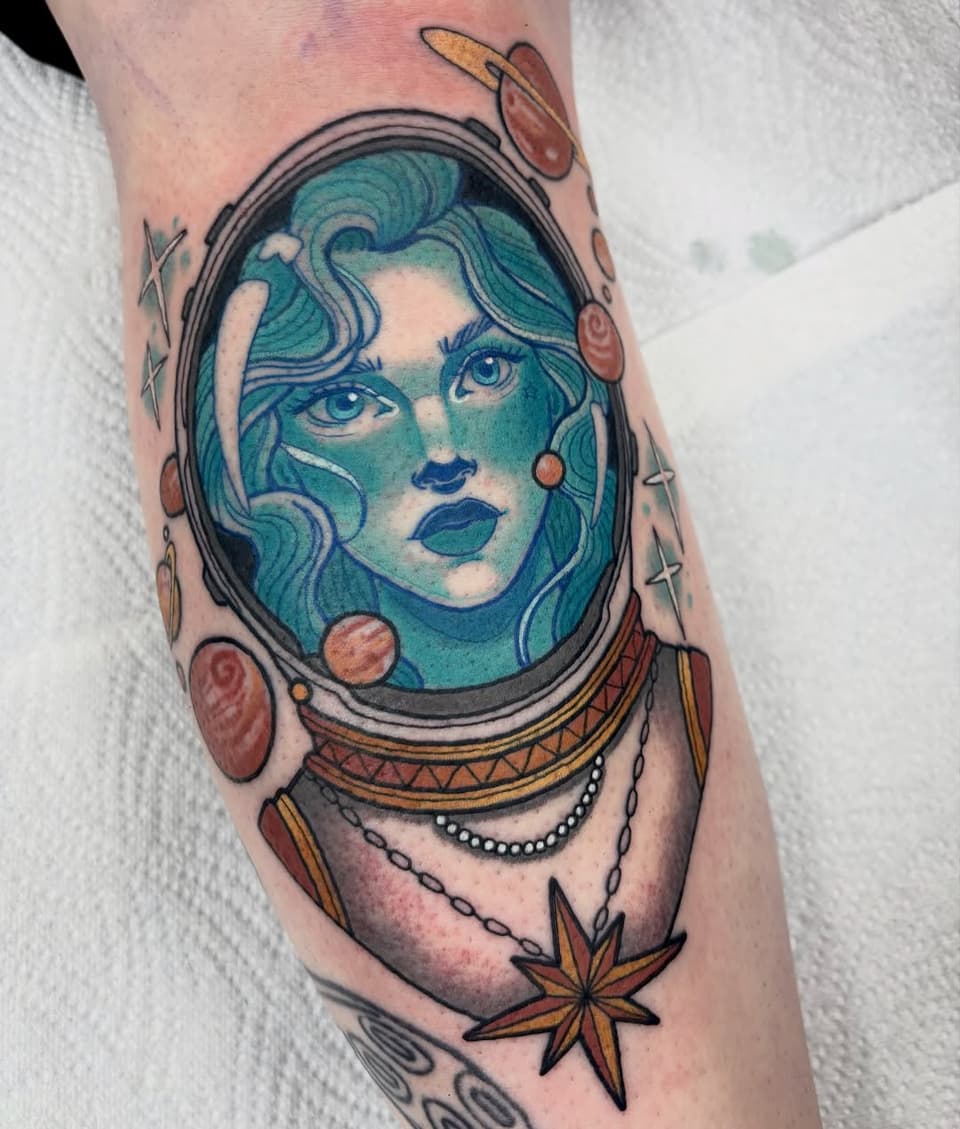 Colorful portrait tattoo of a woman's face with turquoise skin inside a circular frame, surrounded by ornamental elements and stars on the forearm.