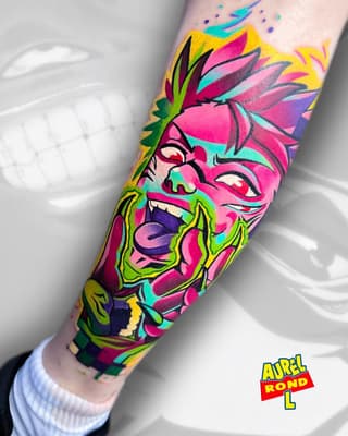 Colorful sleeve tattoo featuring a fierce face with spiky hair, bold outlines, and vibrant magenta, yellow, cyan, and green pigmentation on the forearm.