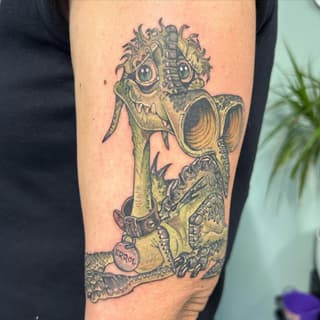 Colorful tattoo on upper arm featuring a grotesque creature with wild hair, large eyes, mechanical elements, and a tag reading "ERROL" in yellow and black ink.