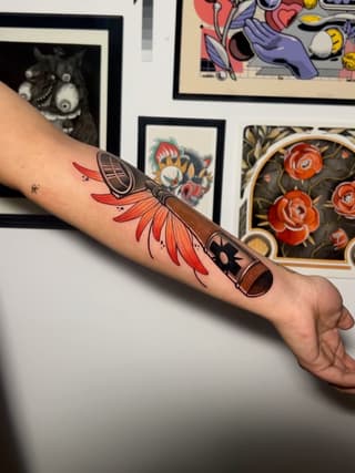 Colorful feather and wooden brush tattoo on forearm featuring red petals, brown details, and black accents against skin.