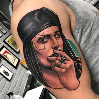 Colored portrait tattoo on upper arm depicting a man with long black hair and head wrap, smoking a cigarette, with green and orange accents.