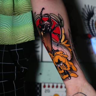 Colorful tattoo on an arm featuring a insect with black body, red circular background, orange and yellow wing details, and decorative floral elements.