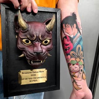 Award-winning demon mask tattoo in burgundy and gold with a separate colorful arm sleeve featuring an anime character, cat figure, and decorative elements.