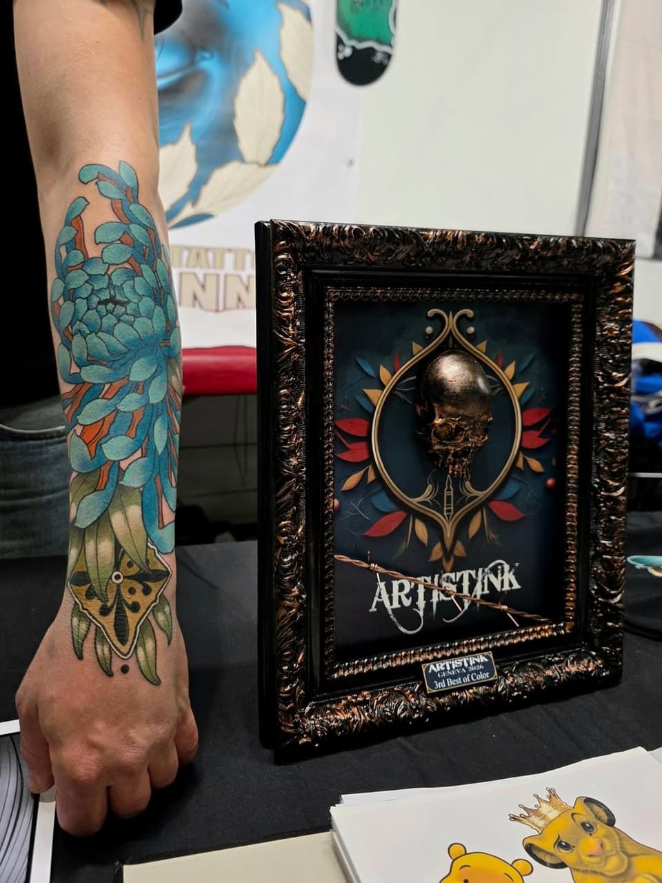 Colorful blue and yellow floral tattoo on forearm featuring layered petals and ornamental details, displayed next to a framed artwork.