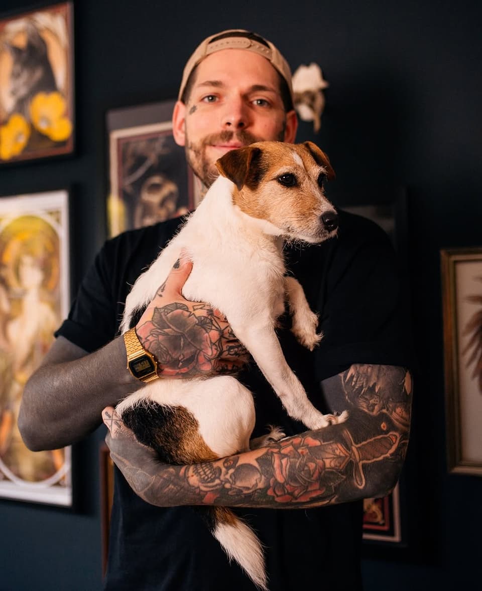 Tattooed man holding small brown and white dog, displaying full-sleeve floral and figurative tattoos in red and black on both arms.