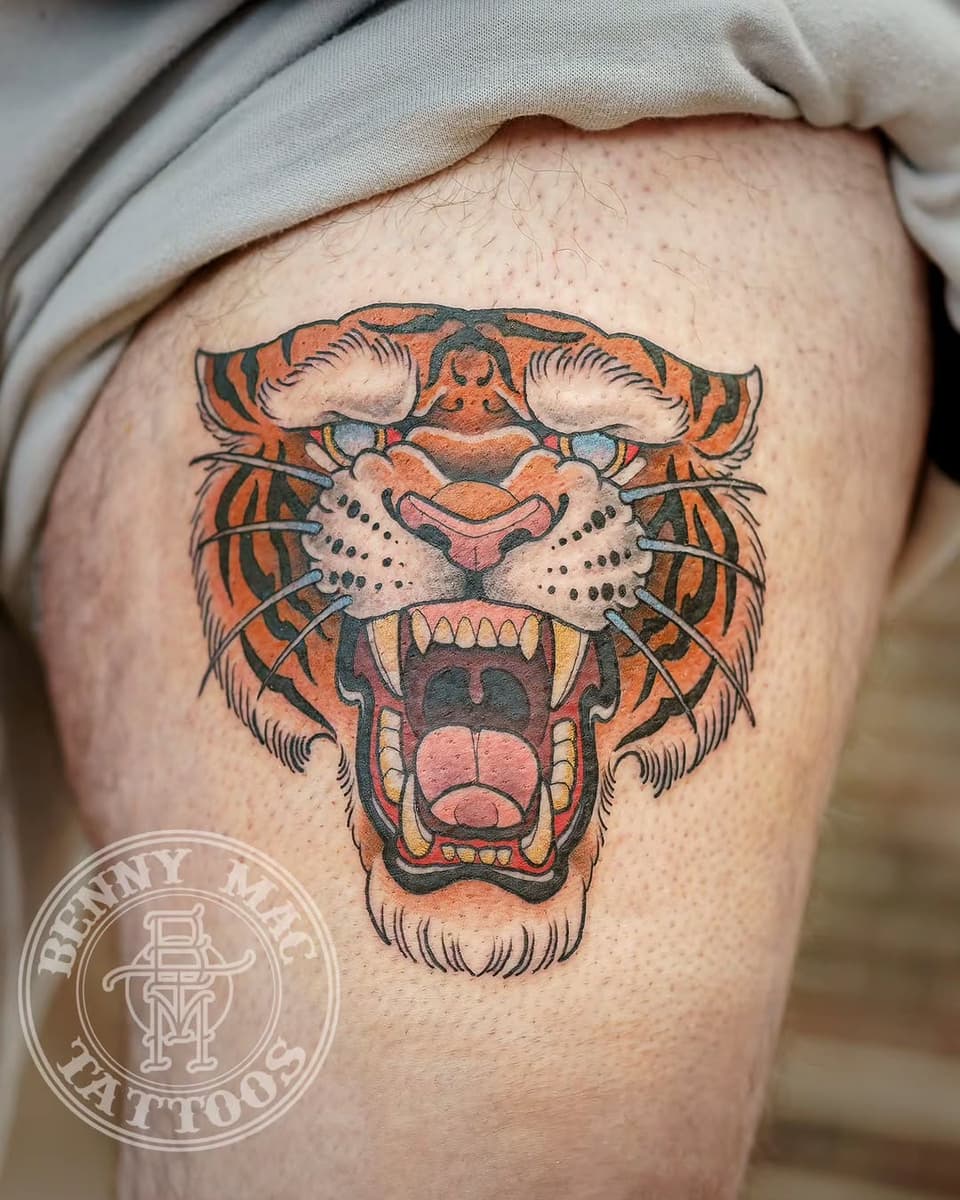 Tiger head tattoo on thigh with orange and black striped markings, open mouth showing fangs and pink interior, positioned on light skin.