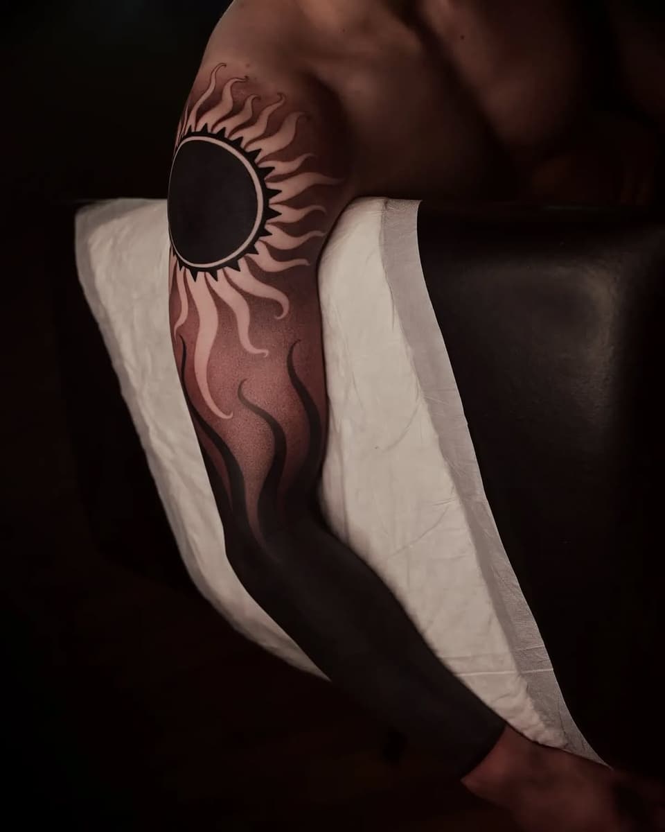 Sun with radiating flame-like rays tattoo on upper arm in brown and black ink, displayed on skin with white gauze wrapping.